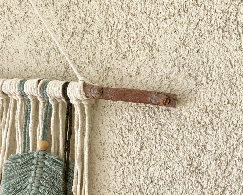 K&ouml;nnte beinhalten: Ein Makramee-Wandbehang mit einer Holzstange und einem Farbspiel aus Hellblau und Wei&szlig;. Der Behang zeigt verschiedene Knoten und Texturen.