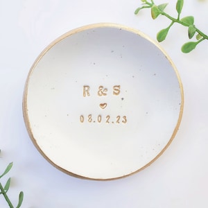 May include: White ceramic ring dish with a gold rim. The dish has the initials "R & S" and the date "08.02.23" inscribed in gold.