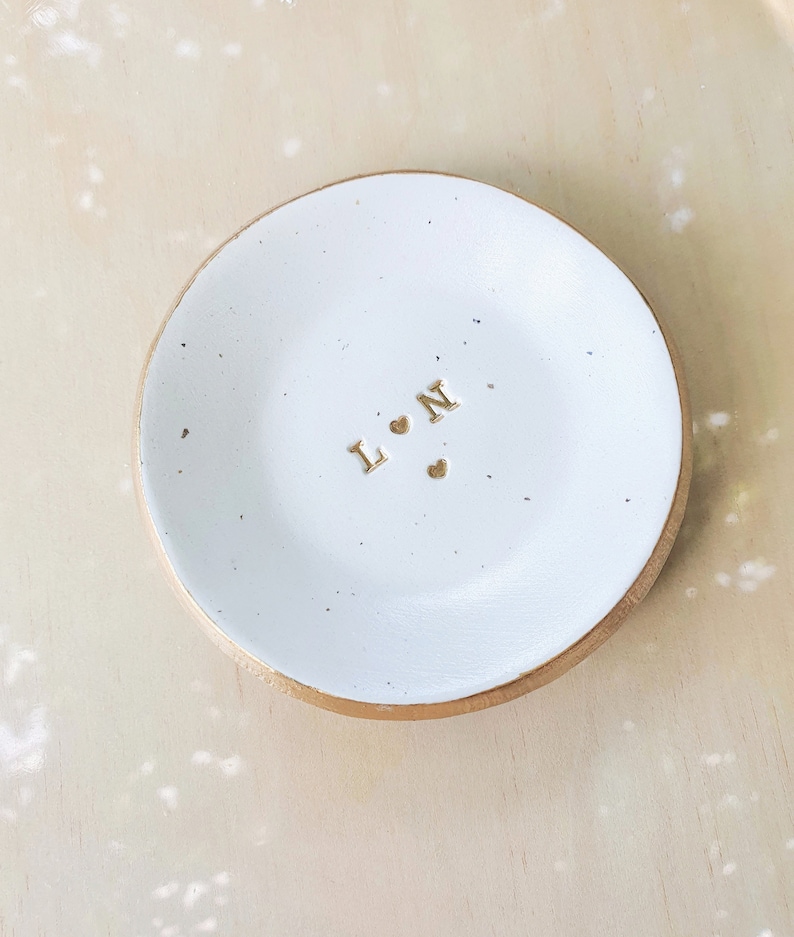 May include: White ceramic ring dish with a gold rim. The dish has the letters "L" and "N" with two small hearts in between.
