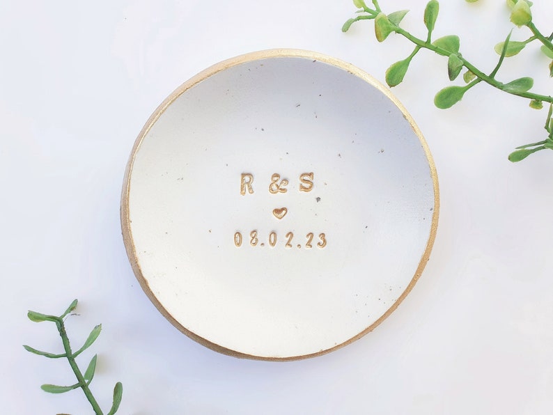 May include: White ceramic ring dish with a gold rim. The dish has the initials "R & S" and the date "08.02.23" inscribed in gold.