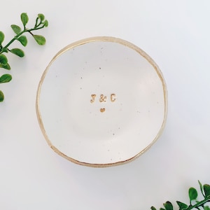 May include: White ceramic ring dish with a gold rim and the initials "J & C" in gold lettering. A small gold heart is centred below the initials.