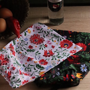 May include: Two fabric pouches with floral patterns, one white with red flowers and the other black with red flowers. A bottle of clear liquid with a label is in the background, along with a basket and some eggs.