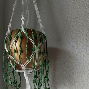 Könnte beinhalten: Ein weißer Makramee-Pflanzenhänger mit grünen Perlen und einem goldenen Topf. Der Hänger hängt an einem Deckenhaken.