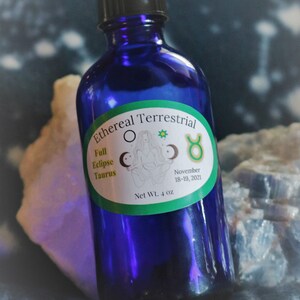 Puede incluir: Una botella de vidrio azul con una tapa negra y una etiqueta verde que dice "Ethereal Terrestrial Full Eclipse Taurus November 18-19, 2021 Net Wt. 4 oz". La etiqueta también presenta una imagen estilizada de una persona en una pose de yoga con una luna creciente y un símbolo de toro.