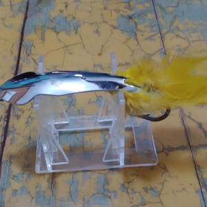 May include: A silver fishing lure with a yellow feather tail. The lure is on a clear plastic display stand.