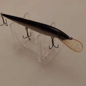 May include: A black and silver fishing lure with a clear plastic tail. The lure is on a display stand with three hooks.