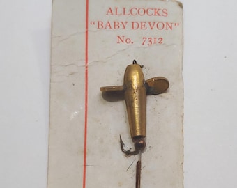 May include: A vintage fishing lure, the "Baby Devon" by Allcocks, is displayed on a worn card. The lure is gold-toned with a propeller design and two hooks. The card has red vertical lines and text that reads "ALLCOCKS BABY DEVON No. 7312".
