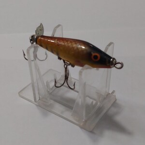 May include: A vintage fishing lure with a propeller and two treble hooks. The lure has a brown and orange body with black accents and an orange eye. It is displayed on a clear plastic stand.