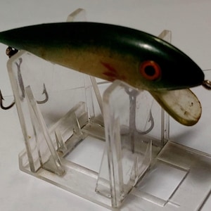 May include: A vintage fishing lure with a dark green top, tan body, and red eye. The lure has a small, white, angled lip and two treble hooks. It is displayed on a clear plastic stand.