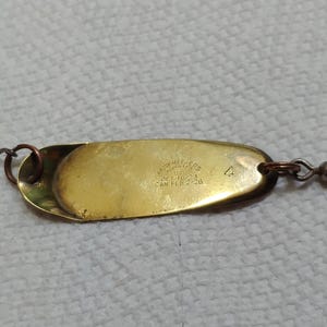 May include: A vintage fishing lure with a gold-colored, oval-shaped body and a silver hook. The lure has a swivel and is designed to attract fish. The body has text that is difficult to read.