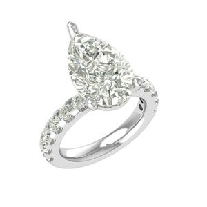 Puede incluir: Un anillo de plata con un diamante en forma de pera como pieza central. La banda está adornada con diamantes redondos más pequeños, creando un efecto brillante. El anillo se presenta sobre un fondo blanco, resaltando su brillo y diseño.