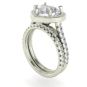 Puede incluir: Un anillo de compromiso plateado con una piedra central en forma de pera y un halo de piedras más pequeñas. La banda se divide en dos, cada una con pequeñas piedras adicionales. El anillo está sobre un fondo blanco.