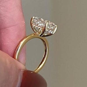 Puede incluir: Un anillo de oro con una gema grande, clara y de talla ovalada. La banda del anillo es de oro pulido, y la gema se sujeta con delicadas garras. El anillo está sujeto por el dedo de una persona.