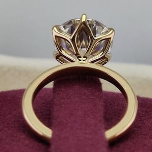 Puede incluir: Un anillo de compromiso de oro con una gema grande y transparente. El anillo presenta un engaste calado único que acuna la piedra. La banda es de oro pulido simple. El anillo se exhibe sobre una superficie burdeos.