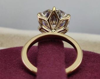 May include: A gold engagement ring with a large, clear gemstone. The ring features a unique, openwork setting that cradles the stone. The band is a simple, polished gold. The ring is displayed on a burgundy surface.