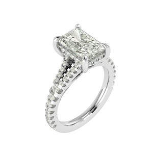 Könnte beinhalten: Ein Verlobungsring mit Diamanten im Radiant-Schliff. Der Ring hat einen großen, rechteckigen Diamanten in einer Krappenfassung. Das Band besteht aus weißem Metall und ist mit kleineren Akzentdiamanten besetzt. Der Ring hat ein geteiltes Schaftdesign.