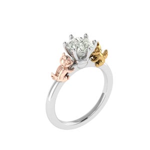 Puede incluir: Un anillo plateado con una gran gema transparente en el centro. Dos pequeñas figuras de dibujos animados en oro rosa y oro flanquean la gema. El anillo está sobre un fondo blanco.