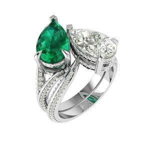 Może przedstawiać: Srebrny pierścionek z dwoma kamieniami szlachetnymi w kształcie gruszki. Jednym z nich jest żywy zielony szmaragd, a drugim przezroczysty, błyszczący diament. Obręcz pierścionka zdobią małe, błyszczące kamienie.