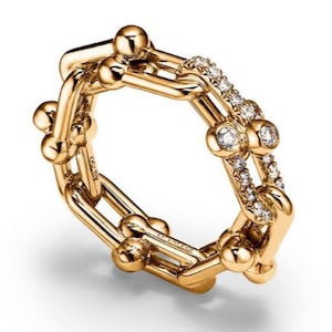 Puede incluir: Un anillo dorado con un diseño de eslabones de cadena. El anillo presenta pequeños acentos redondos y está adornado con una fila de pequeños diamantes brillantes. El anillo es una joya.