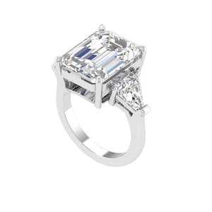 Puede incluir: Un anillo de compromiso de platino con un gran diamante central de talla esmeralda, flanqueado por dos diamantes más pequeños en forma de pera. El anillo tiene una superficie pulida y reflectante.