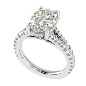 Puede incluir: Un anillo de compromiso de oro blanco con un gran diamante de talla ovalada como pieza central. La banda está adornada con diamantes más pequeños, realzando el brillo y el diseño general del anillo. El anillo está sobre un fondo blanco.