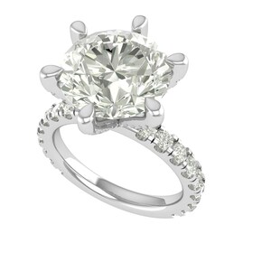 Könnte beinhalten: Ein silberner Verlobungsring mit einem großen, klaren, rund geschliffenen Diamanten als Mittelpunkt. Das Band ist ebenfalls mit kleineren Diamanten besetzt, was einen funkelnden Effekt erzeugt. Der Ring ist vor weißem Hintergrund.