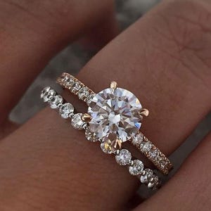 May include: Two rings are shown, a gold band with small diamonds and a larger diamond solitaire ring. The solitaire ring has a large, round, clear diamond. The second ring has a band of small, round diamonds.