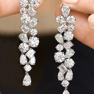 Puede incluir: Elegantes pendientes colgantes con una cascada de brillantes diamantes blancos. Los pendientes presentan una variedad de cortes de diamantes, incluyendo formas redondas, de pera y esmeralda, engastados en una montura de plata u oro blanco. El diseño es simétrico y llamativo.