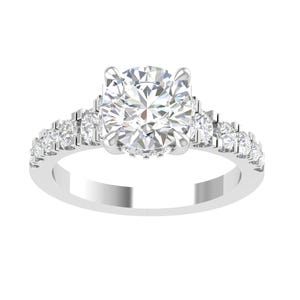 Puede incluir: Un anillo de compromiso plateado con un gran diamante central de talla redonda. Pequeños diamantes adornan la banda y los lados de la piedra central, creando un efecto brillante. El anillo se presenta sobre un fondo blanco.