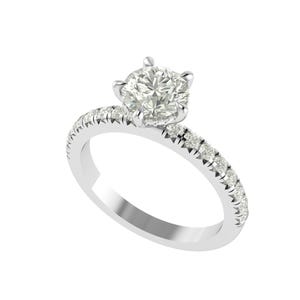 Puede incluir: Un anillo de compromiso plateado con un gran diamante redondo y transparente en el centro. La banda también está adornada con diamantes más pequeños. El anillo se presenta sobre un fondo blanco, resaltando su brillo y diseño.