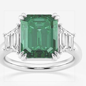 Puede incluir: Un anillo de plata con una gran piedra verde de talla esmeralda. Dos piedras blancas más pequeñas, de talla baguette, flanquean la piedra principal. El anillo tiene un diseño clásico y elegante.