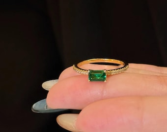 May include: A gold ring with a rectangular emerald-cut green gemstone. The band is gold with small, clear stones. The ring is shown on a person's finger against a black background.