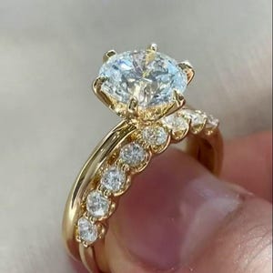 Könnte beinhalten: Ein goldener Verlobungsring mit einem großen, runden, klaren Edelstein als Mittelpunkt. Das Band ist goldfarben, und ein kleinerer Ring mit mehreren kleineren, klaren Edelsteinen befindet sich daneben. Der Ring wird von den Fingern einer Person gehalten.
