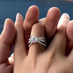 Puede incluir: Un anillo de compromiso plateado con una piedra preciosa grande, redonda y transparente en el centro. El anillo tiene una banda dividida con piedras de acento más pequeñas. El anillo está en el dedo de una persona, sostenido por la mano de otra persona.