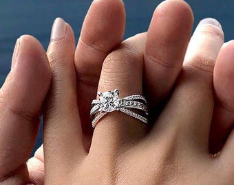 May include: A silver engagement ring with a large, round, clear gemstone at the center. The ring has a split band with smaller accent stones. The ring is on a person's finger, held by another person's hand, with a blurred ocean and mountain background.