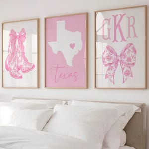 May include: Three framed wall art prints in light pink and white. The left print shows pink cowboy boots with a bow. The center print features a white map of Texas with a heart and the word Texas. The right print displays the letters GKR and a bow.