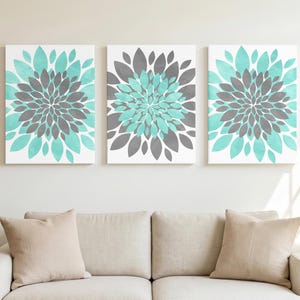 以下が含まれることがあります： 対称的な花の模様が描かれた3枚の長方形のキャンバスプリントセット。各プリントには、鮮やかな白い背景に対して、濃い灰色の花びらの中央のクラスターと、その周囲を囲む明るいターコイズブルーの花びらの層が表現されています。