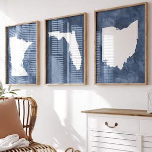 May include: Three framed wall art pieces featuring white state outlines against a blue background. The frames are a light brown color. The art is displayed above a white cabinet with a wooden top. A wicker chair with a brown pillow and white blanket is in the foreground.