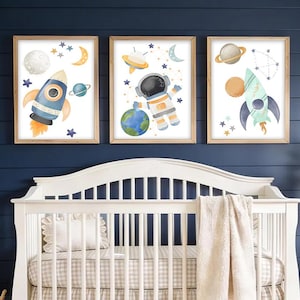 May include: Three framed watercolor art prints featuring space-themed illustrations. The prints depict a rocket, an astronaut, and a rocket with planets and stars. The artwork is displayed above a white crib with a beige and white checkered pattern.
