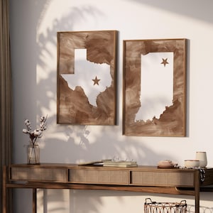 May include: Two framed wall art pieces. One features the state of Texas in white with a brown star, set against a brown, textured background. The other depicts Indiana, also in white with a brown star, with a similar brown background. Both are in wooden frames.