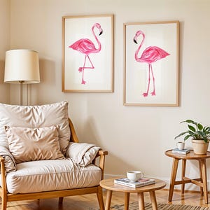 May include: Two framed flamingo art prints. Each print features a pink flamingo with a black beak and long legs, set against a white background. The frames are light-colored wood. The art is displayed in a living room setting with a chair, side tables, and a lamp.