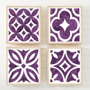 May include: Four framed art prints with geometric patterns in shades of purple and white. Each print features a different design, including floral and diamond shapes, set against a white background. The frames are made of light-coloured wood.