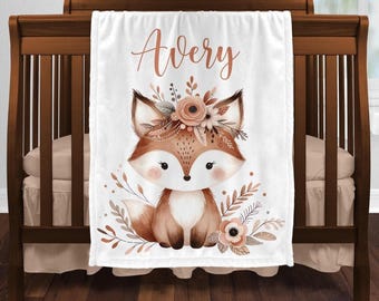 May include: A white fleece blanket featuring a central illustration of a brown fox with a floral crown. The name Avery is printed in a cursive copper-toned font above the fox. The design includes surrounding botanical elements in muted earth tones.
