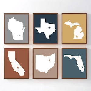 May include: Six framed prints featuring state outlines: Wisconsin, Texas, Michigan, California, Ohio, and Florida. Each state is white against a colored background, with a star marking a city. The frames are brown.