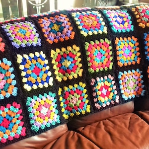 May include: A colorful crocheted throw blanket with a black background and squares of various colors, including red, blue, green, yellow, and orange. The blanket is draped over a brown leather couch.