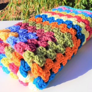 May include: A colorful crocheted blanket with stripes of red, orange, yellow, green, blue, and purple. The blanket is folded in half and the stripes are visible on the outside.