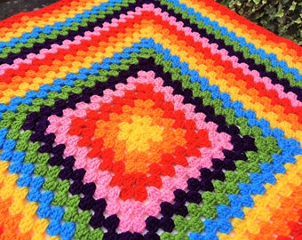 May include: A rainbow crochet blanket with a granny square pattern. The blanket is made with different shades of red, orange, yellow, green, blue, purple, and pink yarn.