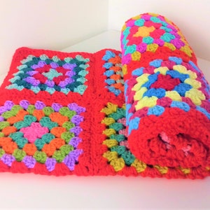 May include: A red crocheted blanket with colorful granny squares. The blanket is rolled up on one side, revealing the colorful squares.