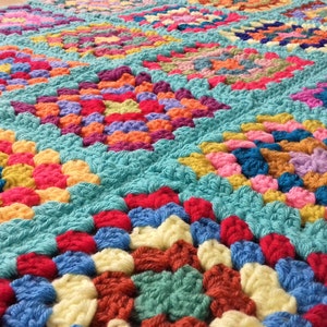 May include: A colourful crocheted blanket featuring a range of bright colours, including red, yellow, blue, green, and purple. The blanket is made up of squares with a textured pattern.
