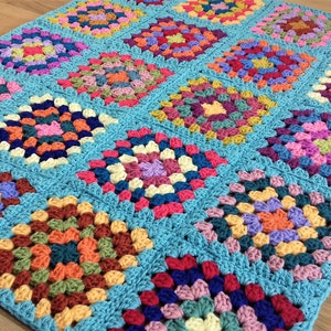 May include: A colourful crocheted blanket made of granny squares in various shades of pink, blue, green, yellow, and orange. The blanket is made of a soft, textured yarn and has a light blue background.
